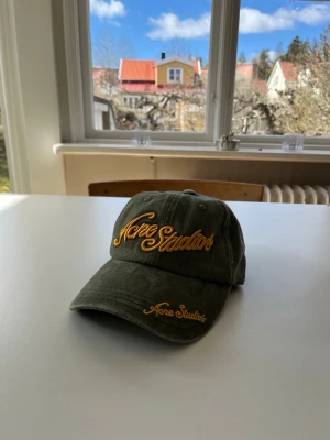 Grön keps från Acne Studios - Säljer en grön keps från Acne Studios med orange broderad logga både på fronten och skärmen. Kepsen har klassisk form med böjd skärm och justerbar baksida. Materialet är mjukt och slitstarkt bomullstyg, perfekt nu inför sommaren. Vid frågor eller funderingar är det bara att säga till Mvh Olof!