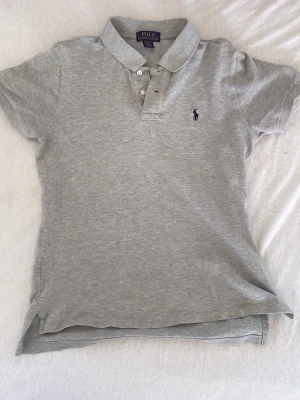 Grå polotopp från Ralph Lauren - Säljer en stilren grå polotopp från Ralph Lauren med klassisk krage och knappar framtill. Toppen har korta ärmar och den ikoniska broderade logon på bröstet. Perfekt för dig som gillar sportiga och tidlösa plagg.