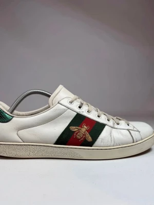 Gucci Ace vita sneakers med bi - Gucci Ace sneakers i vitt läder med klassiska gröna och röda sidoband samt broderad guldfärgad bi. Skorna har vita snören, rund tå och grön detalj vid hälen. Snygg och tidlös modell i premiumläder som sticker ut med sina ikoniska detaljer. Stl 44 passar 45