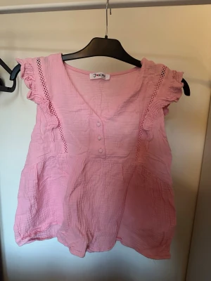 Rosa blus med volang och spets - Supersöt rosa blus från J'aime med volangdetaljer på axlarna, spetsinslag och v-ringning med knappar framtill. Blusen är ärmlös och har en luftig, lätt struktur i bomull. Perfekt för en fräsch och somrig look!