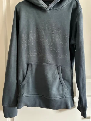 Mörkgrå hoodie med tryck - Snygg mörkgrå hoodie från Replay med diskret tryck framtill. Klassisk känguruficka och ribbade muddar vid ärmslut och nederkant. Perfekt för en avslappnad stil och enkel att matcha med jeans eller joggers.