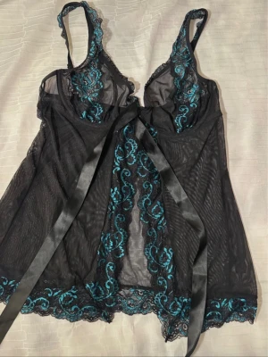 Svart set för kvinnor från Obsessive st. L/XL - Säljer en set  med turkosa broderade spetsdetaljer från Obsessive. Västen har snygg snörning framtill med ett brett satinband och vackra spetskanter längs nederdelen och axelbanden. Perfekt för dig som gillar unika detaljer och elegant design.Polyamid 96%, elastan 4%