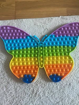 Stor regnbågsfärgad pop it fjäril - Säljer en stor pop it i form av en fjäril med regnbågsfärger. Den har lila, blå, gröna, gula och orangea rader och är tillverkad i mjukt silikon. Perfekt för att stressa ner eller bara ha kul med att trycka på bubblorna.