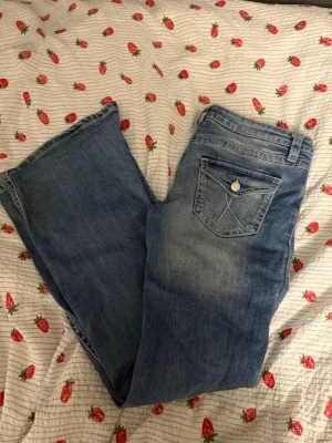Blå bootcut jeans med broderad ficka - Säljer ett par ljusblå bootcut jeans med snygg broderad bakficka och klassiska detaljer. Jeansen har en avslappnad passform och är tillverkade i denim. Har några slitningar men går nog att fixa enkelt därav priset