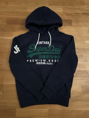Mörkblå Superdry hoodie med tryck - Mörkblå hoodie från Superdry med stor logga och japanska tecken på bröstet. Vit text på ärmen och vita snören i huvan. Klassisk passform med ribbade muddar och mjukt material, perfekt för en avslappnad look.