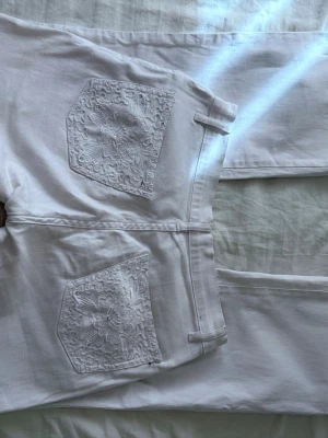 Lågmidjade jeans med detaljer - Fina vita jeans med broderidetaljer och stenar på bakfickorna Liten slits nedtill och stretchigt material  Midja tvärs över: 35 cm Innerbenslängd: 73 cm