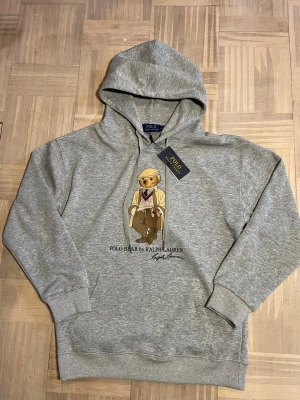 Grå hoodie Polo Bear Ralph Lauren - Säljer en grå hoodie från Ralph Lauren med Polo Bear-motiv på bröstet. Hoodien har huva, ribbade muddar och en klassisk känguruficka. Materialet är mjukt och bekvämt, perfekt för en avslappnad stil. Märket syns tydligt både på taggen och trycket.