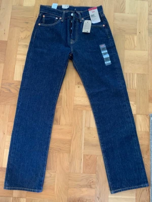 Levi's 501 mörkblå jeans - Klassiska Levi's 501 jeans i mörkblå denim med raka ben och femficksdesign. Jeansens material är 100% bomull och har ikonisk läderpatch bak samt röda Levi's-tag på bakfickan. Perfekt för dig som gillar tidlös stil och kvalitetsjeans.