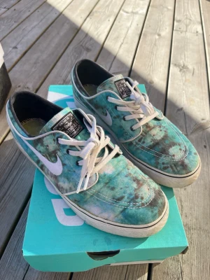 Nike SB Stefan Janoski Tie-Dye - Unika Nike SB Stefan Janoski sneakers med tie-dye mönster i turkos, vitt och brunt. Skorna har vita snören, vit sula och klassisk låg siluett. Materialet är canvas och de har den ikoniska Swoosh-loggan på sidan. Perfekta för dig som vill sticka ut med din stil.