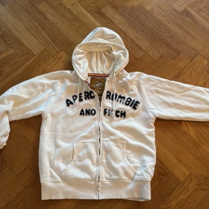 Vit hoodie Abercrombie & Fitch XL - Vit hoodie från Abercrombie & Fitch i storlek XL med broderad logga på bröstet. Tröjan har huva med snörning, dragkedja framtill och två fickor. Mjuk bomullsmix som är skön att bära. Perfekt för chill dagar och streetwear-stil.