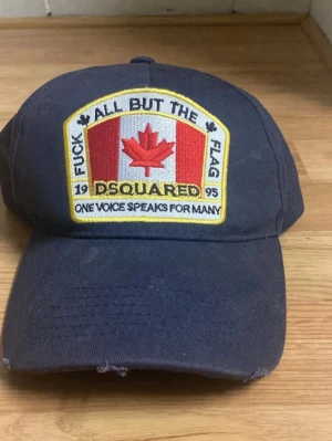 Svart Dsquared keps med patch - Cool svart keps från Dsquared med stor broderad patch framtill som har kanadensisk flagga och text. Kepsen har böjd skärm och justerbar baksida. Materialet är bomull och designen är klassisk med en edgy touch.