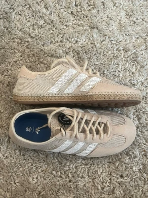 adidas gazelle x clot skor  - Säljer ett par beige adidas x clot skor. Skorna har en låg profil och är tillverkade i en kombination av mocka och textil, vilket ger en snygg och avslappnad look. Perfekta för dig som gillar stilrena och bekväma sneakers. skicket är 10/10 (aldrig använda) kommer me box och blå snören