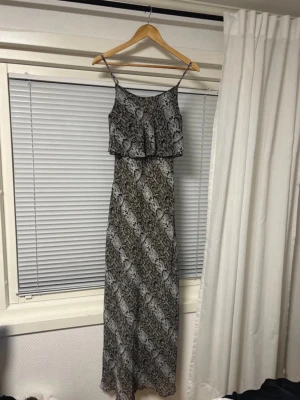 Långklänning med snakeprint - Snygg långklänning med smala axelband och coolt svart och grått snake-mönster. Klänningen har ett lager upptill som ger en avslappnad look och är perfekt för sommarens alla tillfällen. Materialet känns lätt och luftigt.