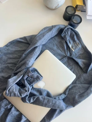 Grå skjorta från Essentials - Säljer en stilren grå skjorta från H&M Essentials. Skjortan har klassisk krage, knappar framtill och långa ärmar med manschett. Materialet är mjukt och bekvämt, perfekt för dig som gillar en enkel och tidlös look.