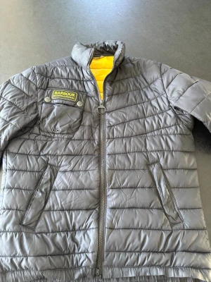 Svart dunjacka från Barbour International herr S - Säljer en svart dunjacka från Barbour International med quiltad design och gul innerfoder. Jackan har hög krage, två snedställda fickor med dragkedja och en bröstficka med Barbour-logga. Perfekt för kalla vinterdagar eller vårdagar. Fläckfri, den enda skavanken är på innerfickan, syns ej utåt. 