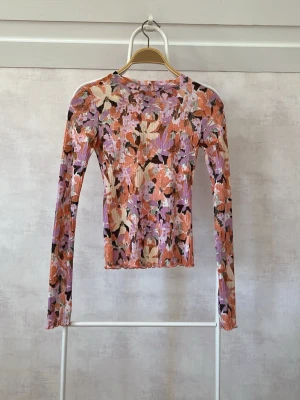 Blommig långärmad topp i ribbad stil - Trendig långärmad topp med blommigt mönster i lila, orange, beige och svart. Ribbad struktur ger en snygg och bekväm passform. Perfekt för dig som vill sticka ut med färg och mönster. Tillverkad i mjuk polyester.
