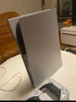 PlayStation 5 med två DualSense-kontroller - Säljer en PlayStation 5 med två vita och svarta DualSense-kontroller. Konsolen har en futuristisk design med vita paneler och svart mittdel. Perfekt för gaming och upplevelser i toppklass. Materialet är plast med en matt och blank finish.