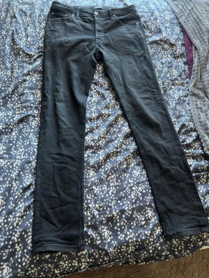 Jack & Jones Svarta raka Jeans Comfort Mike model W30 L34 - Säljer ett par svarta jeans med rak passform och klassisk femficksdesign. Jeansen har normal midja, bälteshällor och stängs med knapp och dragkedja. Materialet är troligtvis bomull med lite stretch för extra komfort. Storleken är W30 L34 