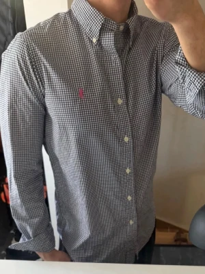 Rutig skjorta från Polo Ralph Lauren - Säljer en klassisk rutig skjorta från Polo Ralph Lauren med svart och vit småmönstrad design. Skjortan har långärmad passform, knappar framtill och en rosa broderad logga på bröstet. Perfekt för dig som gillar stilrena och tidlösa plagg.
