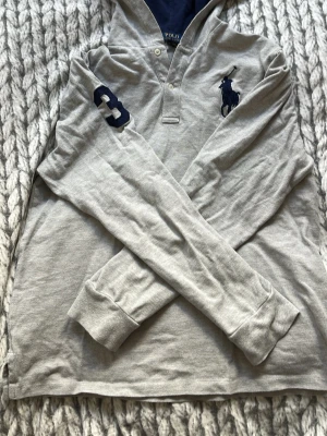Grå hoodie från Polo Ralph Lauren - Snygg grå hoodie från Polo Ralph Lauren med stor mörkblå logga på bröstet och en broderad siffra 3 på ärmen. Tröjan har huva, långa ärmar och ribbade muddar. Tillverkad i mjuk bomull för en avslappnad och sportig look. XL barnstorlek passar som S