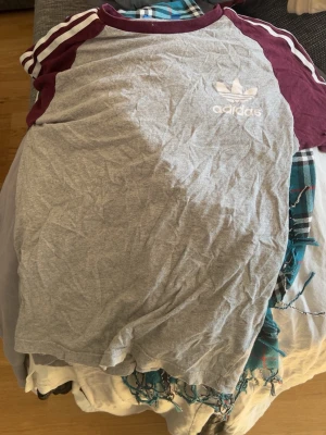 Grå Adidas t-shirt med vinröda detaljer - Säljer en grå t-shirt från Adidas med vinröda raglanärmar och vita ränder på axlarna. Klassisk Adidas-logga på bröstet. Skön passform och tillverkad i bomull, perfekt för dig som gillar sportig stil.