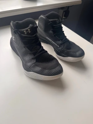 Svarta basket skor från Under Armour - Säljer ett par svarta basket skor från Under Armour med vit sula och logga på sidan. Skorna har högt skaft, snörning och är tillverkade i syntetmaterial med ventilerande mesh och perforeringar för extra komfort. Perfekta för träning och sport.