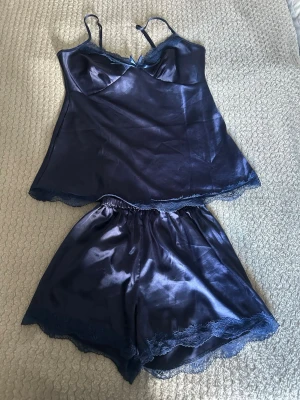 Mörkblå satin nattlinne och shorts med spets - Elegant mörkblått nattlinne och short i satin med tunna justerbara axelband och dekorativ spets vid halsen och stretchig söm i midjan på shortsen. Materialet är glansigt och mjukt, perfekt för en lyxig känsla. Liten rosett framtill ger en extra detalj.