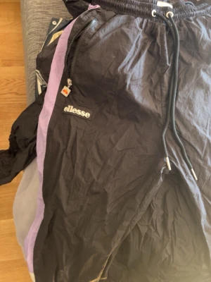 Ellesse svarta tränings byxor  med lila detaljer - Säljer ett par svarta mjukisbyxor från Ellesse med lila sidopaneler och dragsko i midjan. Byxorna har dragkedjeförsedda fickor och Ellesse-logga på vänster ben. Materialet är lätt och har en sportig känsla, perfekt för chill eller träning.