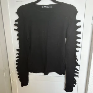 Svart långärmad tröja FB Sister - Svart långärmad tröja från FB Sister med unika utskurna detaljer längs ärmarna som ger en edgy vibe. Tröjan är i mjukt stickat material och har en rund halsringning. Perfekt för dig som vill sticka ut med din stil.