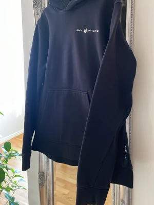 Svart hoodie från Sail Racing  - Svart hoodie från Sail Racing med logga på bröstet och ett diskret tryck på huvan. Klassisk passform, stor framficka och ribbade muddar. Materialet är mjukt och bekvämt, perfekt för chill dagar. Huva med snörning och stilren design.