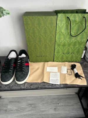 Gucci ace skor  - Snygga Gucci sneakers i svart canvas med klassiskt GG-monogram, svarta skosnören och vit sula. På sidan finns den ikoniska röda och gröna randen. Kommer med originalkartong, dustbag och extra skosnören. Perfekta för dig som gillar exklusiv streetstyle.
