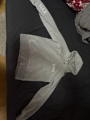 Ljusgrå hoodie från ZYN, Storlek S/XS - Ljusgrå hoodie från ZYN med vit logga på bröstet och huvan. Klassisk känguruficka framtill och dragsko i huvan. Mjuk bomullsblandning och ribbade muddar vid ärmslut och nederkant. Perfekt för en chill och avslappnad stil.