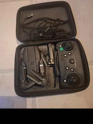 Drönare med handkontroll  - Säljer en kompakt svart drönare med hopfällbara armar och tillhörande fjärrkontroll. Drönaren kommer i en praktisk väska med laddningskabel och har en modern design i plast. Perfekt för dig som vill testa flygning första gången.