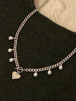 Silverfärgat halsband med pärlor och hjärta - Snyggt silverfärgat rostfritt halsband med chunky kedja, dekorerat med små vita pärlor och ett hjärtformat hänge. Halsbandet har en toggle-låsning framtill som ger en trendig touch. Perfekt accessoar för att lyfta din outfit.