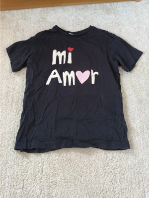 Svart pyjamastopp med text mi Amor - Mysig svart pyjamastopp från KappAhl med trycket 'mi Amor' i vitt och rosa, där bokstaven 'o' är utbytt mot ett hjärta. Rund halsringning och korta ärmar. Perfekt för chilliga kvällar eller lata morgnar. I storlek 146-152/XS. 