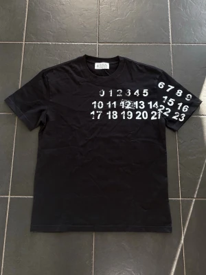 Svart t-shirt med siffror från Maison Margiela - Cool svart t-shirt från Maison Margiela med vit siffror-print över bröstet och ena ärmen. Klassisk rund hals och korta ärmar. Materialet är mjuk bomull och passformen är normal. Perfekt för dig som gillar unika designer och streetstyle.