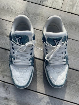 Louis Vuitton vita och blå sneakers - Snygga Louis Vuitton sneakers i vitt och blått med LV-logga på tungan och ovansidan. Skorna har en låg profil, vita skosnören och detaljer i denimliknande material. Perfekta för dig som vill sticka ut med exklusiva sneakers.
