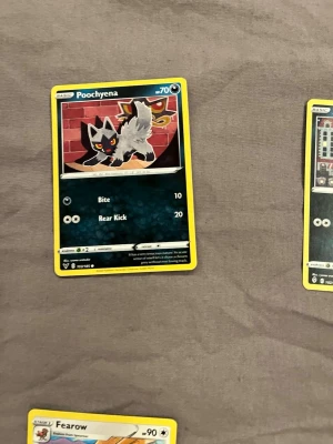 Pokémonkort Poochyena - Säljer ett Pokémonkort med Poochyena-motiv. Kortet har en svart och grå tecknad hund mot en röd tegelvägg, med gula och vita detaljer. Kortet har gula kanter och visar attacker som Bite och Rear Kick. Perfekt för samlare eller Pokémonfans.