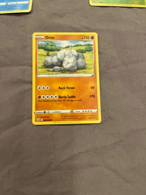 Pokémonkort Onix - Säljer ett Pokémonkort med Onix-motivet. Kortet är orange med en bild på Onix i en naturmiljö. Det är tillverkat av papper och har detaljer som attacknamn och HP-värde. Perfekt för samlare eller Pokémon-fans.
