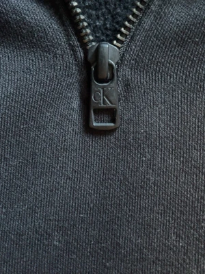 Svart hoodie från Calvin Klein Jeans - Svart hoodie med halv dragkedja och stor CK-logga på bröstet. Hoodien har en huva, ribbade muddar och en stor magficka. Materialet är mjukt och bekvämt, perfekt för chill dagar. Snygg och enkel design med Calvin Klein Jeans-märke.