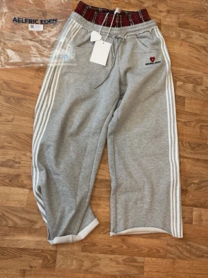 Grå mjukisbyxor Aelfric Eden - HELT NYA, bara testade! Grå mjukisbyxor från Aelfric Eden med vita ränder längs sidorna och en unik röd- och svartrutigt resår i midjan. Byxorna har inbyggda flanellshorts och ett litet hjärta med text på vänster sida. Perfekt för chill och streetwear. (säljer då de var för långa) 