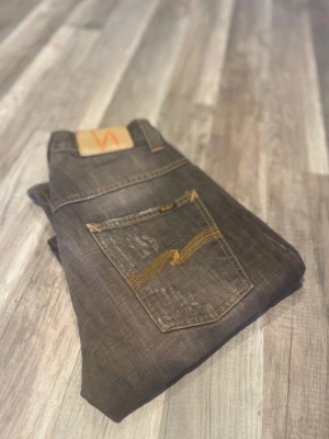 Nudie Jeans mörkgrå denim - Säljer ett par mörkgrå jeans från Nudie Jeans med klassisk gul söm på bakfickorna och läderpatch med logga. Jeansen har rak passform och är tillverkade i slitstark denim. Perfekt för dig som gillar en stilren och cool look.