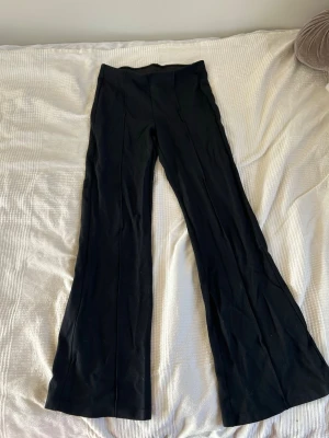 Svarta utsvängda byxor - Säljer ett par svarta utsvängda byxor. Byxorna har en enkel och stilren design utan synliga fickor eller detaljer. Materialet ser ut att vara stretchigt och mjukt, vilket gör dem bekväma att bära. Perfekta för dig som gillar en trendig och clean look.