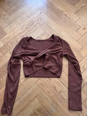 Brun croppad omlottopp från SHEIN - Snygg brun croppad omlottopp från SHEIN i ribbad polyester. Toppen har långa ärmar och en twistad omlottdetalj framtill som ger en trendig look. Perfekt att styla med höga jeans eller kjol.