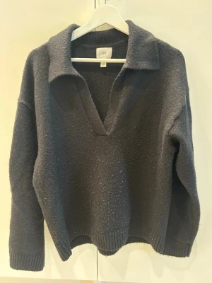 Mörkgrå stickad tröja med krage - Mysig mörkgrå stickad tröja från H&M med djup v-ringning och bred krage. Tröjan har långa ärmar och ribbade muddar vid ärmslut och nederkant. Perfekt för lager på lager och enkel att styla till jeans eller kjol.