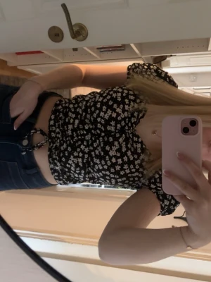 Topp - Söt croppad topp från H&M med svart botten och vita blommor. Toppen har korta puffärmar, v-ringning och snörning framtill. Perfekt för sommarens alla tillfällen och ger en trendig vibe till din outfit.