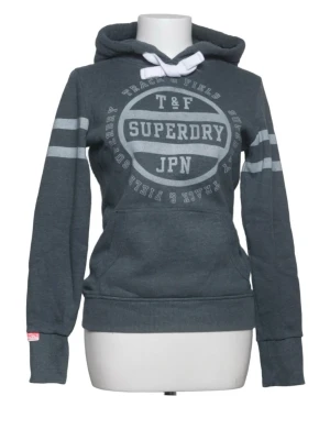 Mörkgrå hoodie från Superdry - Mörkgrå hoodie från Superdry med vit logga och text på bröstet. Har huva med vita dragsnören, känguruficka och ljusgråa ränder på ärmarna. Tillverkad i mjukt bomullsmaterial och har ribbade muddar vid ärmslut och nederkant.
