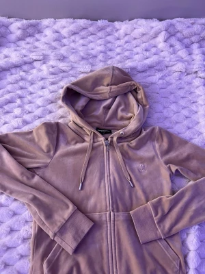 Ljuslila velour hoodie från Juicy Couture - Säljer en ljuslila hoodie i mjuk velour från Juicy Couture. Hoodien har dragkedja framtill, fickor på sidorna, justerbar huva med snören och ett diskret Juicy Couture-märke broderat på bröstet. Perfekt för en chill och trendig look.