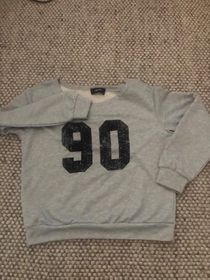 Grå  one shoulder sweatshirt med tryck från Cider - Grå sweatshirt med svart tryck av siffran 90 på bröstet. Tröjan har rund halsringning, långa ärmar och ribbade muddar vid ärmslut och nederkant. Perfekt för en avslappnad och sportig stil.