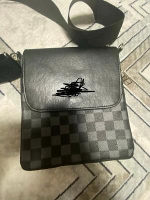 Svart axelväska med rutigt mönster - Snygg svart axelväska med bred axelrem och klassiskt rutigt mönster i grått och svart. Väskan har lock och dragkedja på sidan, perfekt för att bära det viktigaste. Materialet ser ut att vara konstläder och textil.